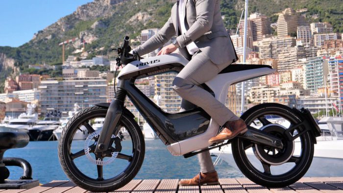 A model in a tan suit, at a dock, trying out the Novus One Electric Motorcycle with a lake in the background